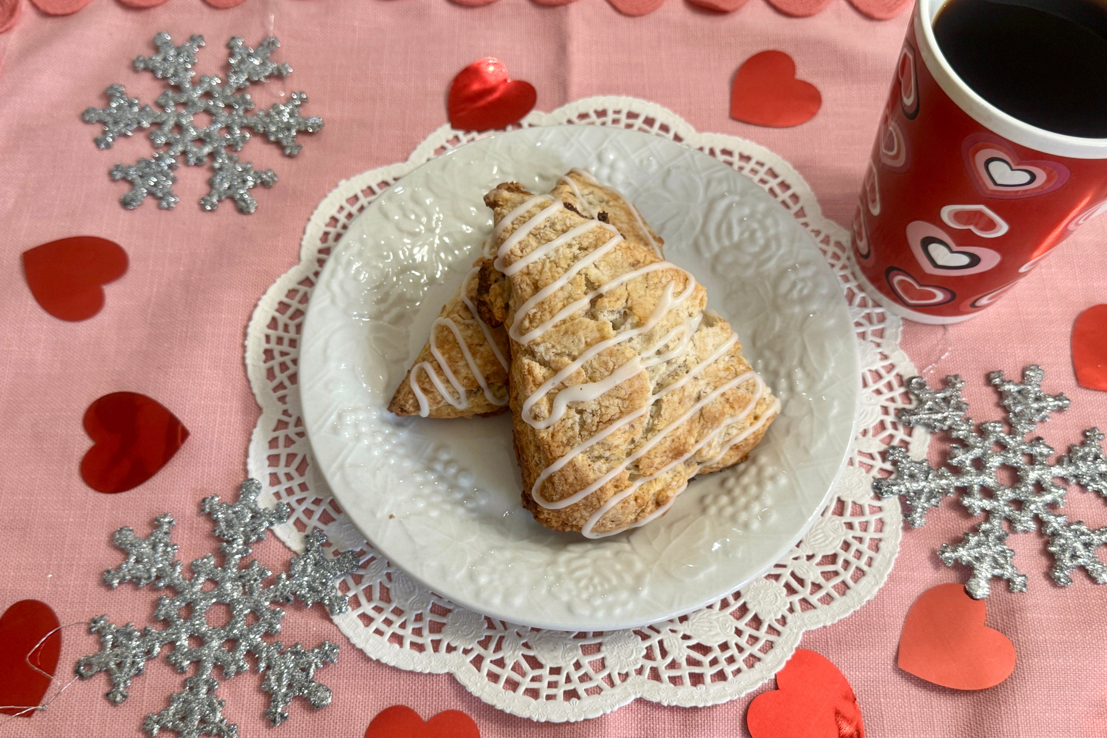 Two strawberry scones on a white plate with a red mug, surrounded by decorative hearts and snowflakes on a pink background.